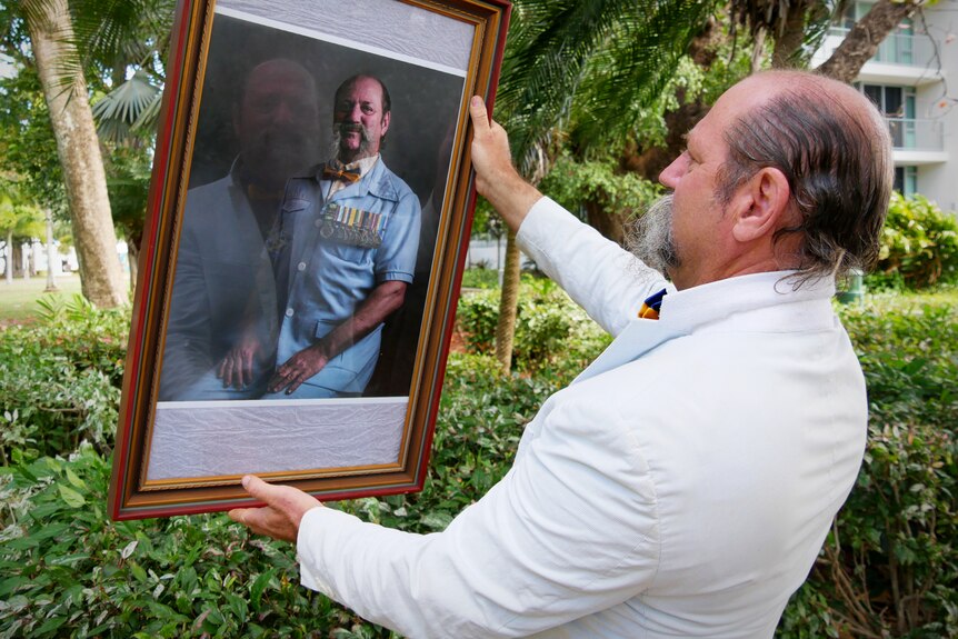 A man holds up and examines a framed photo of himself wearing Army medals