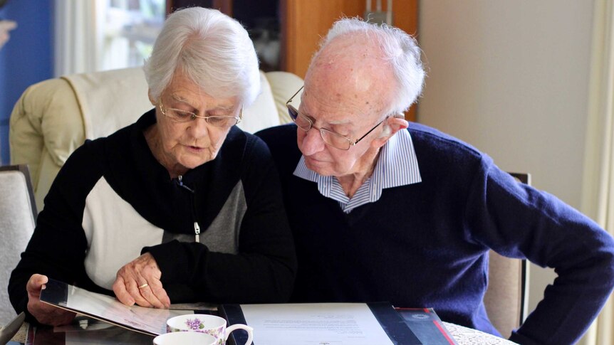An elderly couple sit next to each other drinking tea and flipping through an album.