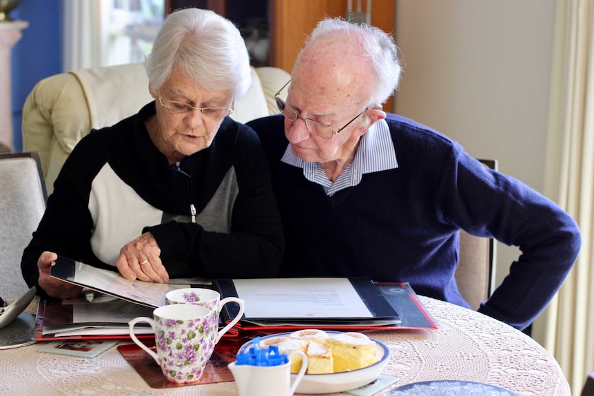 An elderly couple sit next to each other drinking tea and flipping through an album.