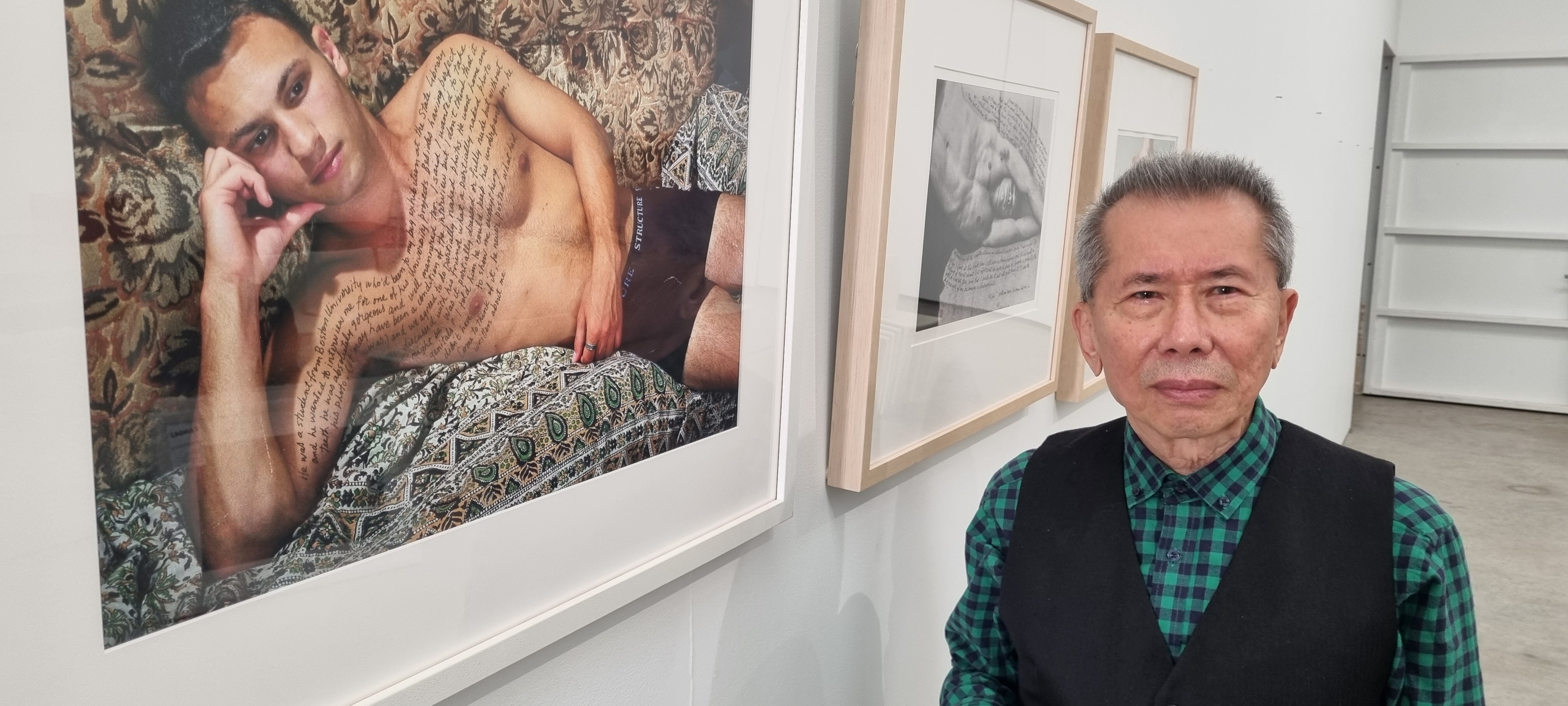 Photographer William Yang in September 2022, beside a photo of a man without a shirt on and writing on his chest