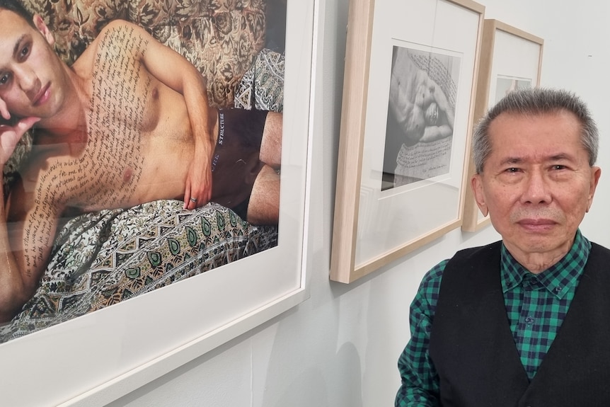 Photographer William Yang in September 2022, beside a photo of a man without a shirt on and writing on his chest