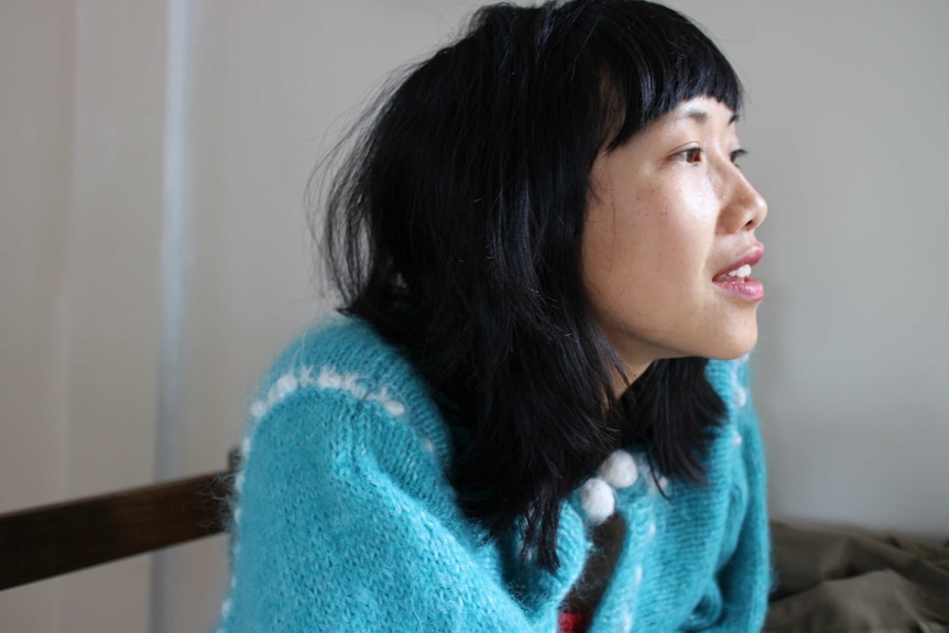 The playwright, wearing a light blue jumper and a cropped bob haircut, stares out of a window against a white wall.