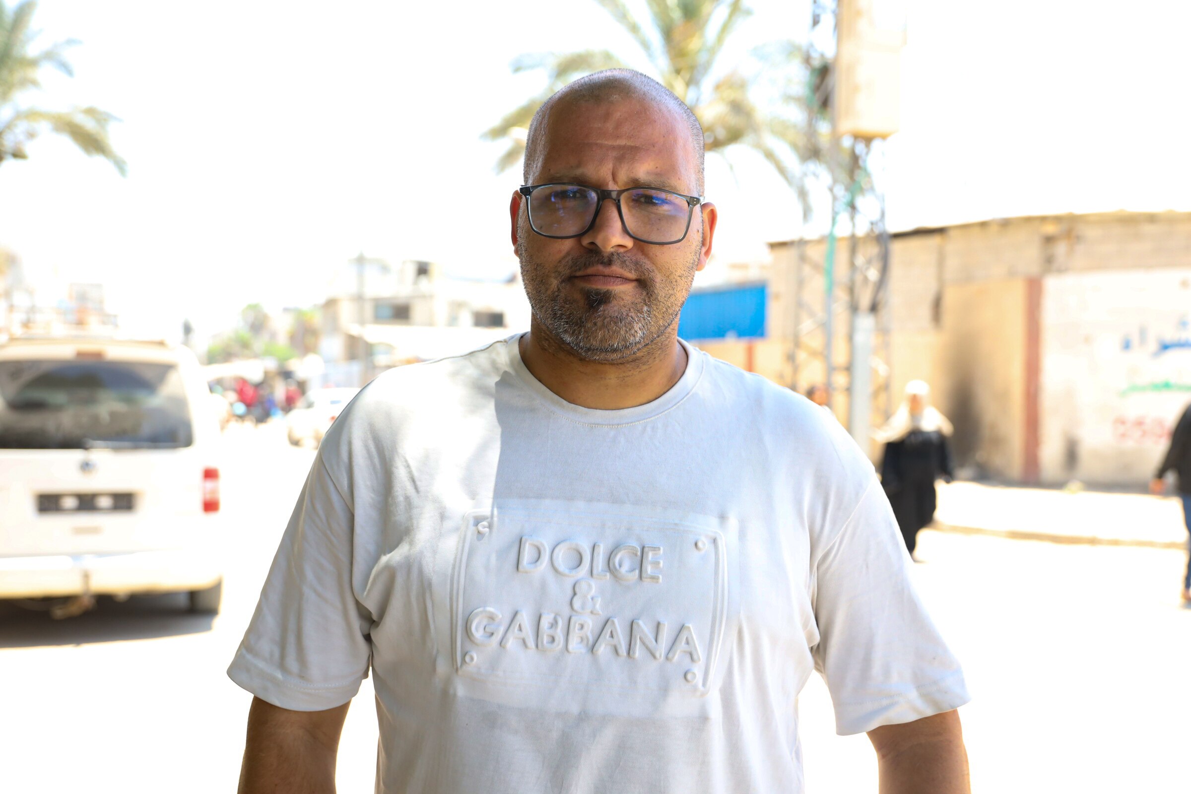 Mohammed Skiek wears spectacles and a white t shirt as he stands near a road and looks into the camera.