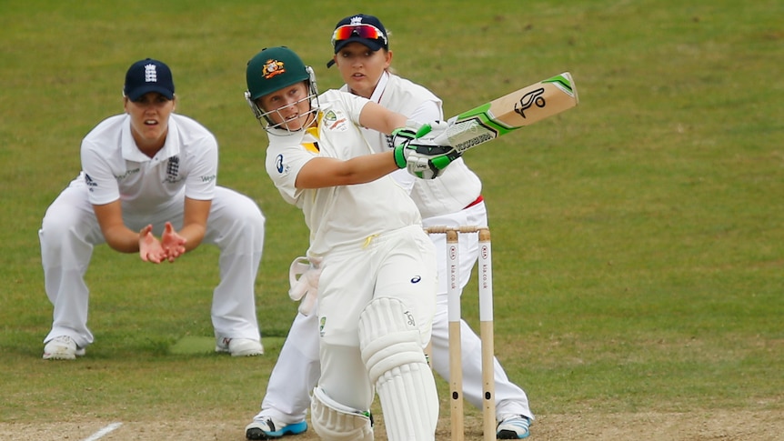 A cricket player swings her bat after hitting a red ball away from the two opposition players in shot.
