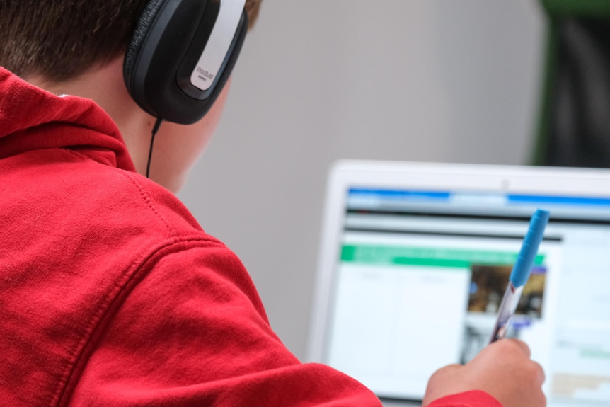 A young person sits in front of a computer screen wearing headphones and taking notes