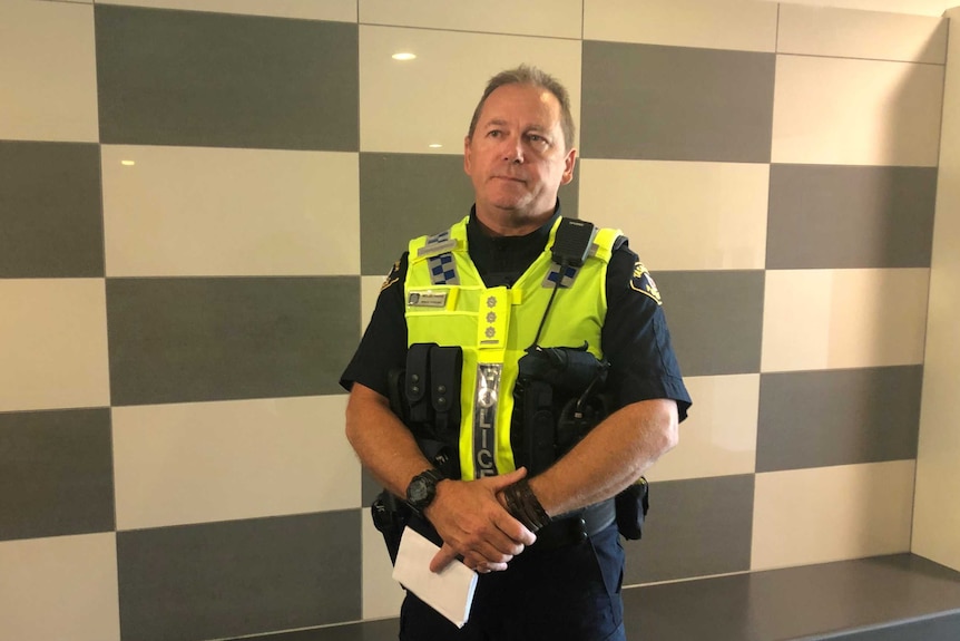 A police officer in a uniform and hi vis vest stands with hands folded in front of him.