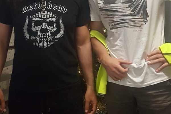 Two men stand side by side for a photograph. one of them wears a metal head shirt, the other has a vest around his hips.