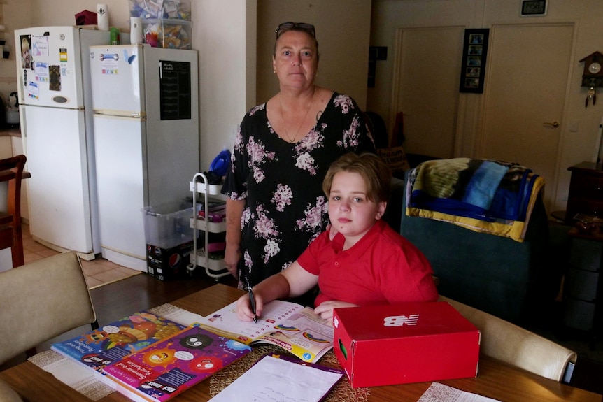 woman stands behind child who sits at kitchen table with books