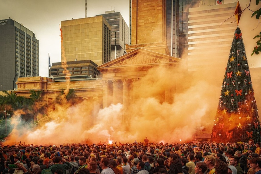 Orange flare smoke drifts past a large building and over a crowd of people standing near a Christmas tree