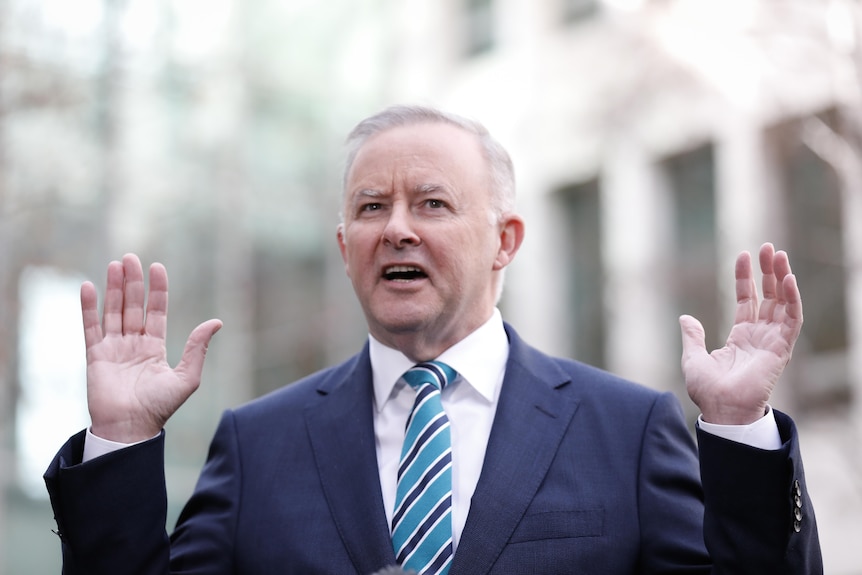 A man in a blue tie and suit puts his hands up in the air as he talks.