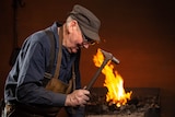 A man leans in with a hammer in his hand. In the background are the flames of a fire.