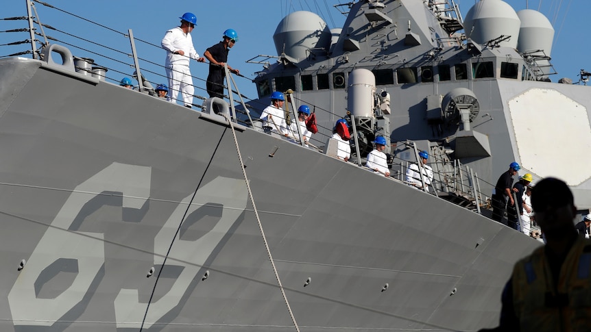 A side view of a grey US warship with the number 69 on side and white clothed sailors on deck