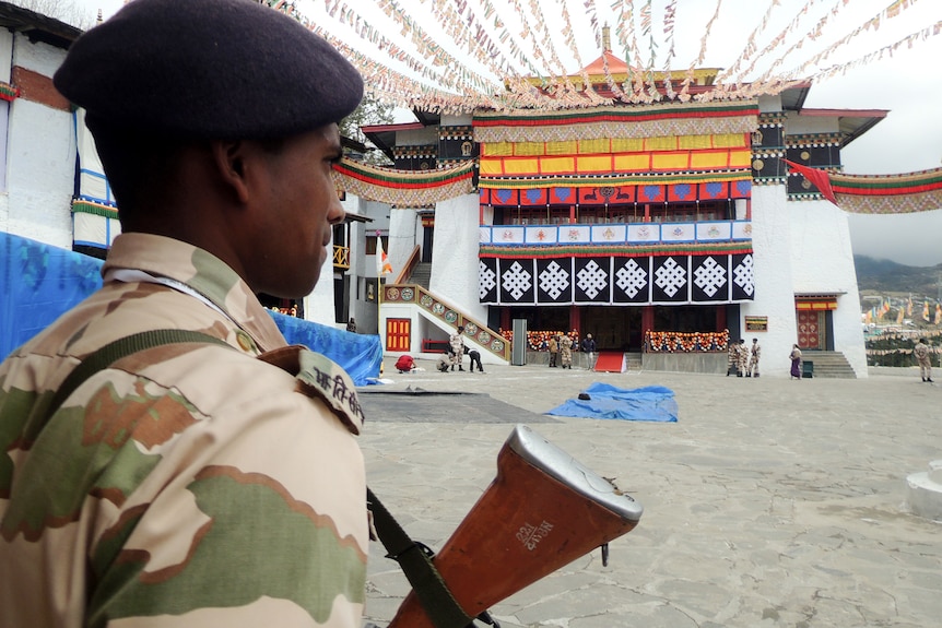 An Indian soldier wearing a hat and army uniform stands on guard with a gun outside a white building.