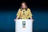 A woman wearing a yellow and green dress stands at a lectern to give a speech during an event