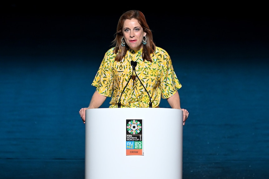 A woman wearing a yellow and green dress stands at a lectern to give a speech during an event