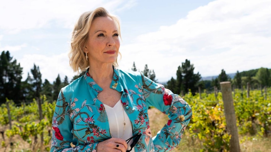A woman with blonde hair and a blue, patterned shirt looks over vineyards. 