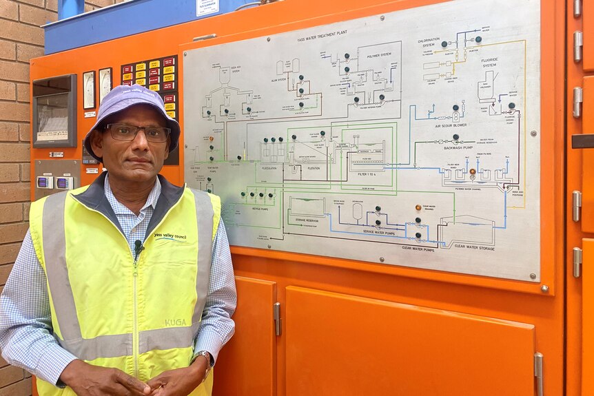 A man in a yellow high-vis vest stands in front of a switchboard for a water filtration plant.