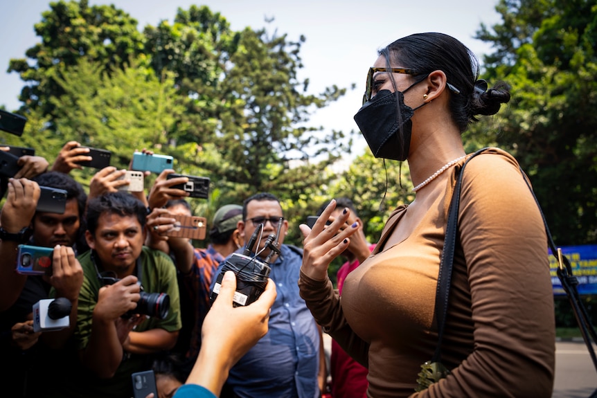 A woman wearing a black face mask and sunglasses, photographed from the side, speaks to a media pack