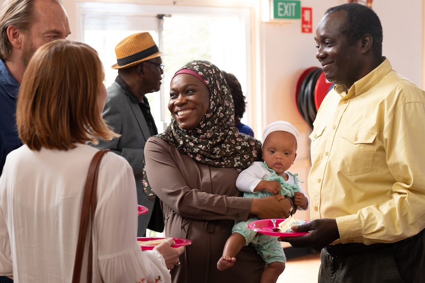 Bolude Watson and Andrew Luri in the film Hearts and Bones, Bolude is holding a baby and they're at an event