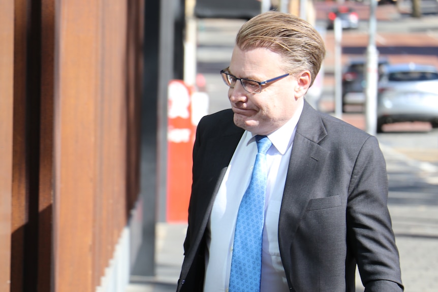 A close shot of WA Ombudsman Chris Field walking into a building wearing spectacles, a dark grey suit, blue tie and white shirt.