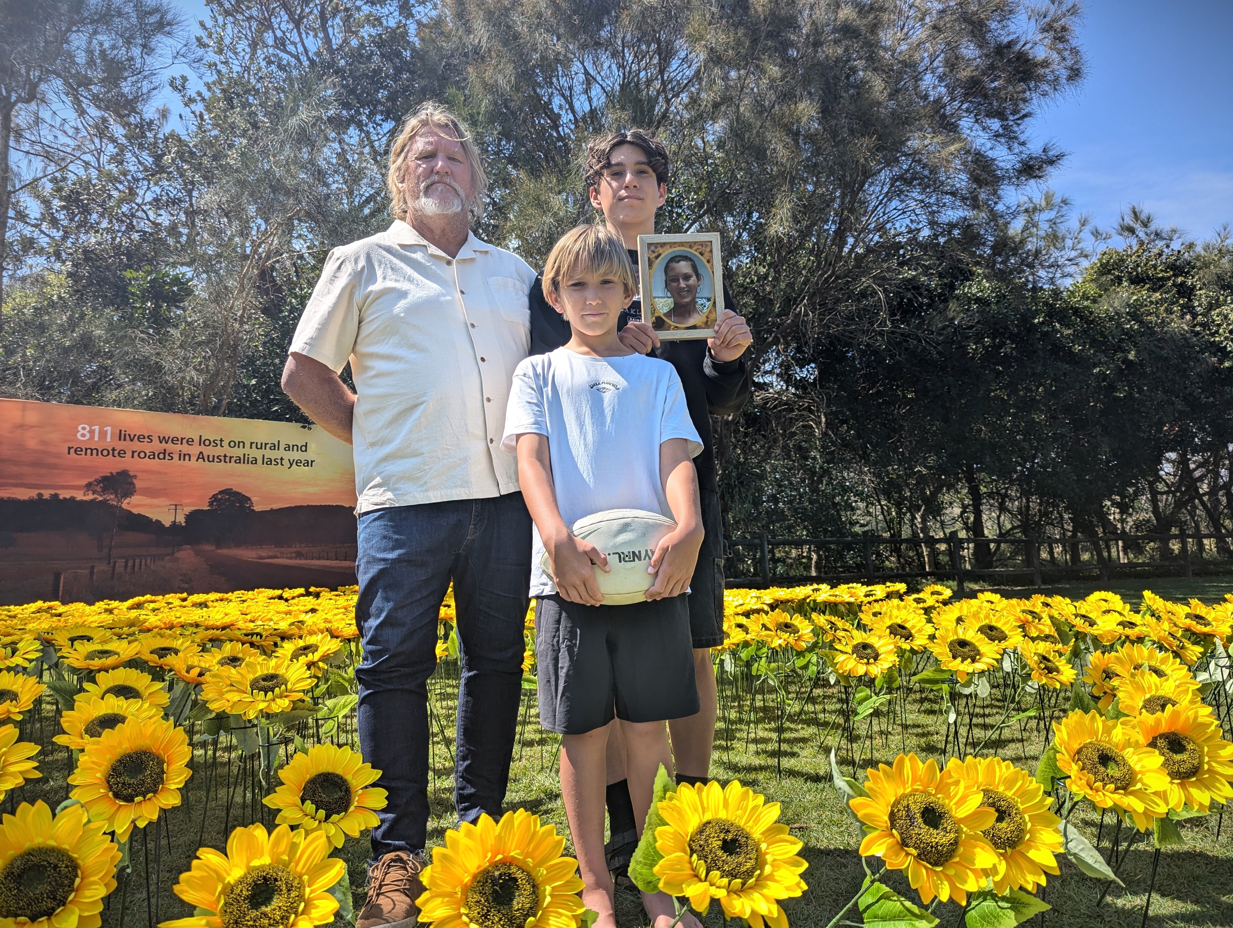 A man and two boys stand with a football and photo frame in the midst of massed sunflowers in front of a billboard