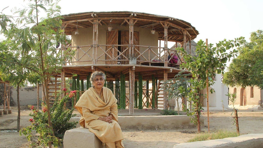 A woman sits on a concrete block in front of a bamboo and mud structure she designed.