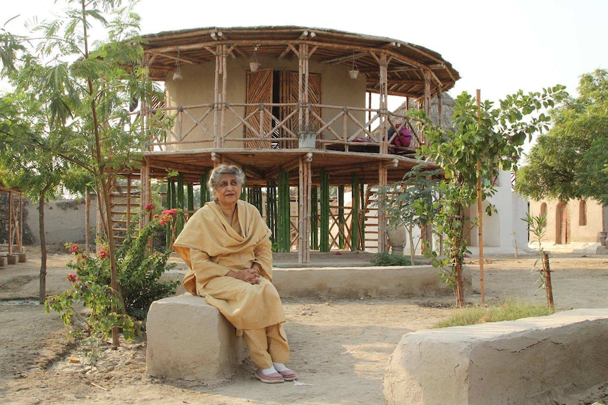 A woman sits on a concrete block in front of a bamboo and mud structure she designed.