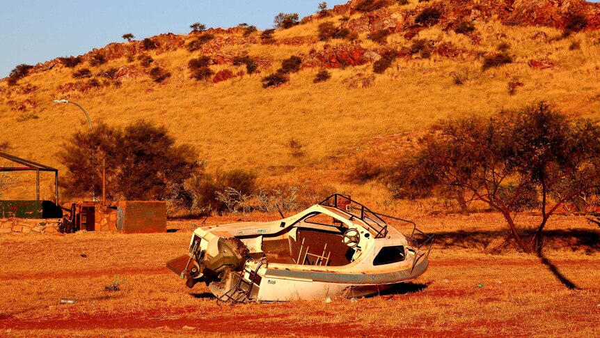 A wrecked motor boat lies on a dry piece of scrubland next to a couple of trees.