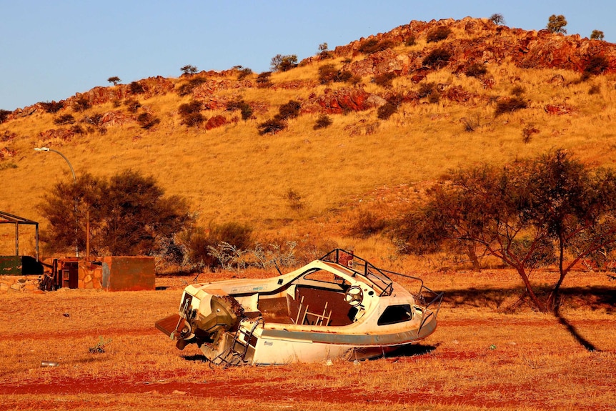 A wrecked motor boat lies on a dry piece of scrubland next to a couple of trees.