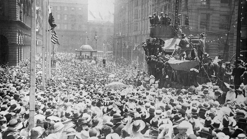 A crowd of people gathered in Martin Place. Military figures are addressing the crowd from a replica sailing ship.