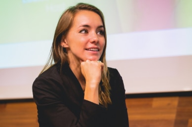 Brunette woman wearing black suit sits cross-legged on a grey couch holding microphone