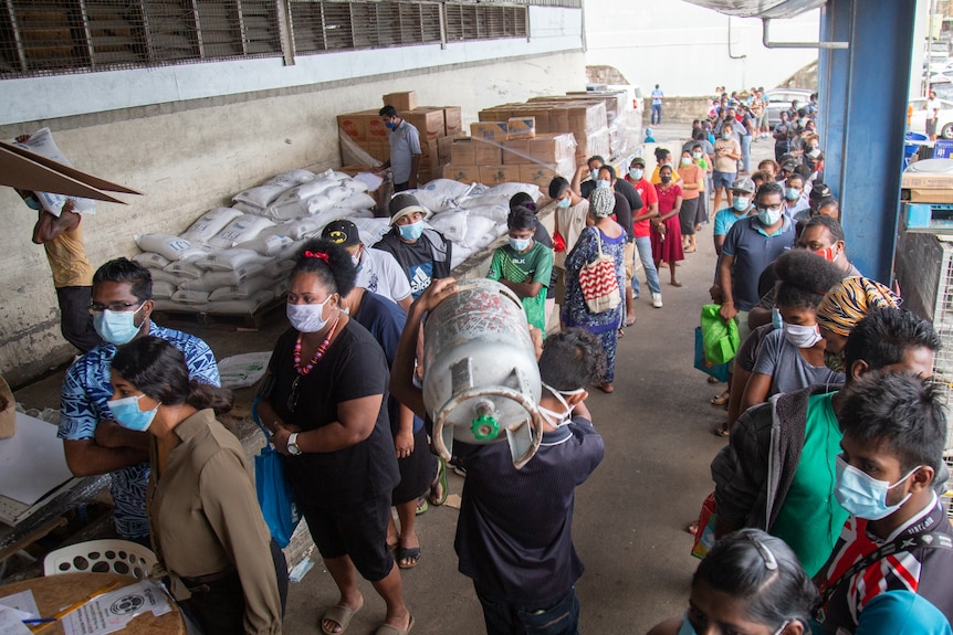 Long lines of people in masks in front of bags of rice. 