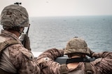 Soldiers in fatigues look out off the deck of a ship, one using a walkie talkie and another using binoculars