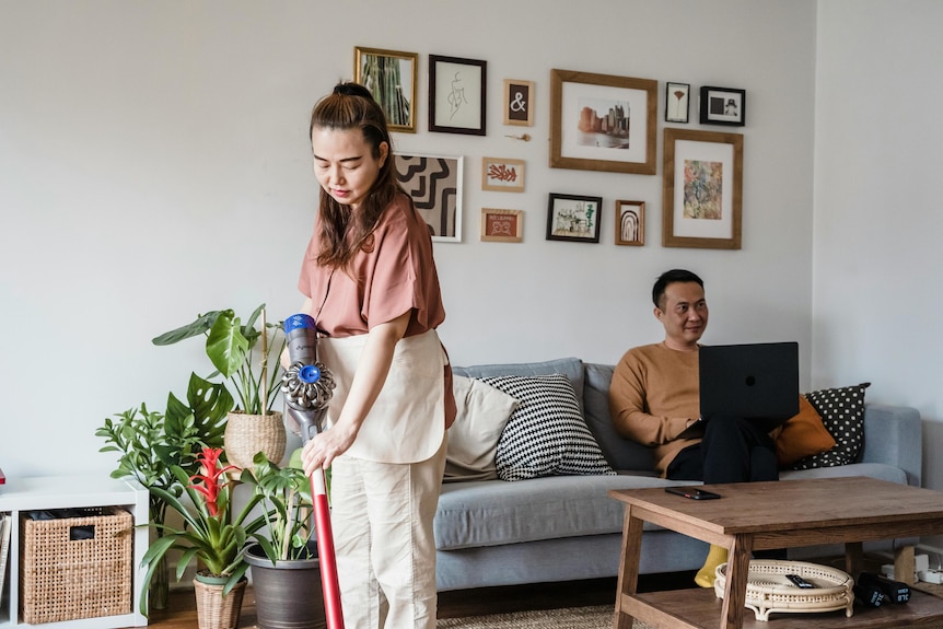 A woman vacuuming the floors while a man sits on a couch and uses a laptop. The living room has plants and prints on the wall. 