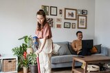 A woman vacuuming the floors while a man sits on a couch and uses a laptop. The living room has plants and prints on the wall. 