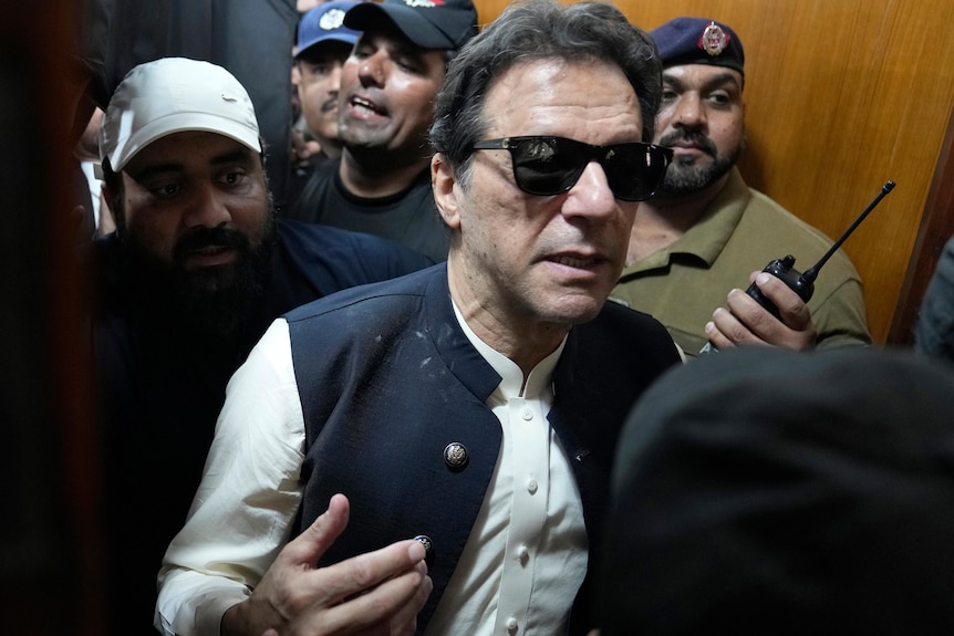 Imran Khan walks through a dense crowd of men as he tries to leave a courtroom while wearing sunglasses.