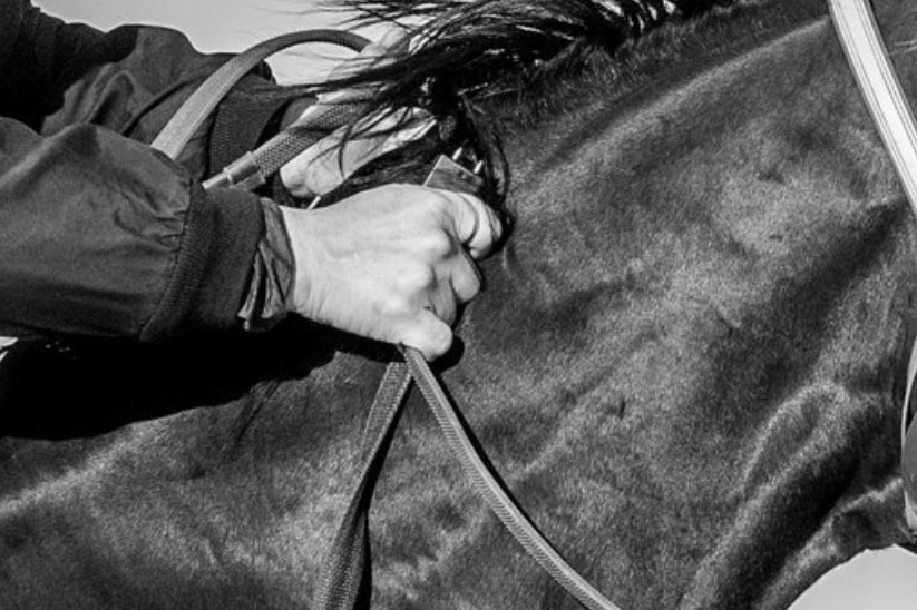 A close up of a jockey's hand holding a small electrical device in his fist as he grips the reins on a horse.