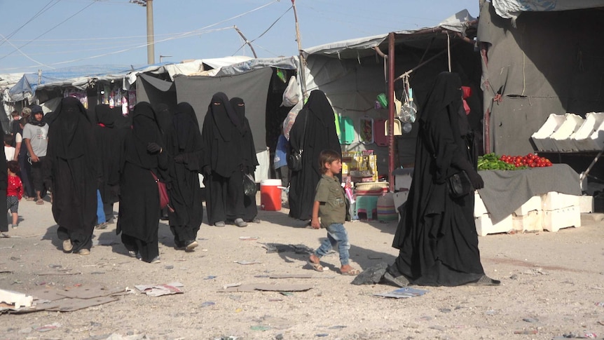 Child in Syrian refugee camp walks among women in black billowing robes.