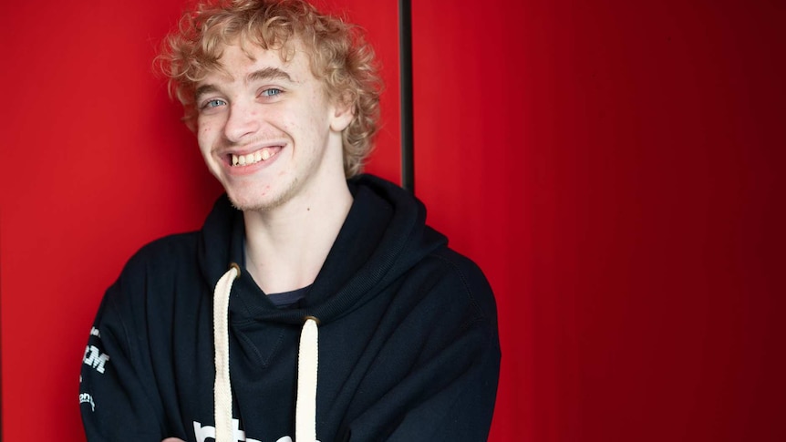 A young man in a black jumper with his arms crossed stands in front of a red wall.