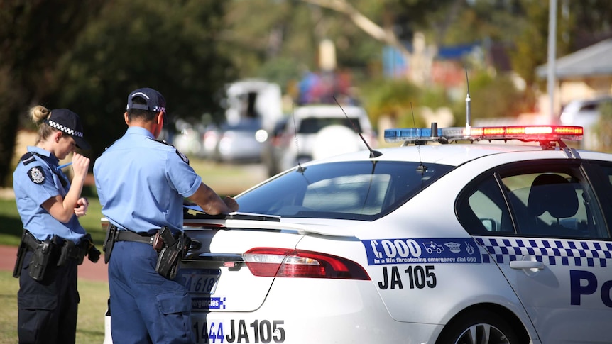 Two police officers stand at the rear of a police car with paperwork.