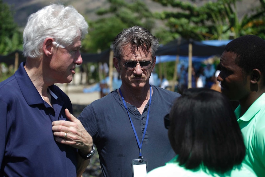 Bill Clinton and Sean Penn, wearing sunglasses and a lanyard, speak to some locals.