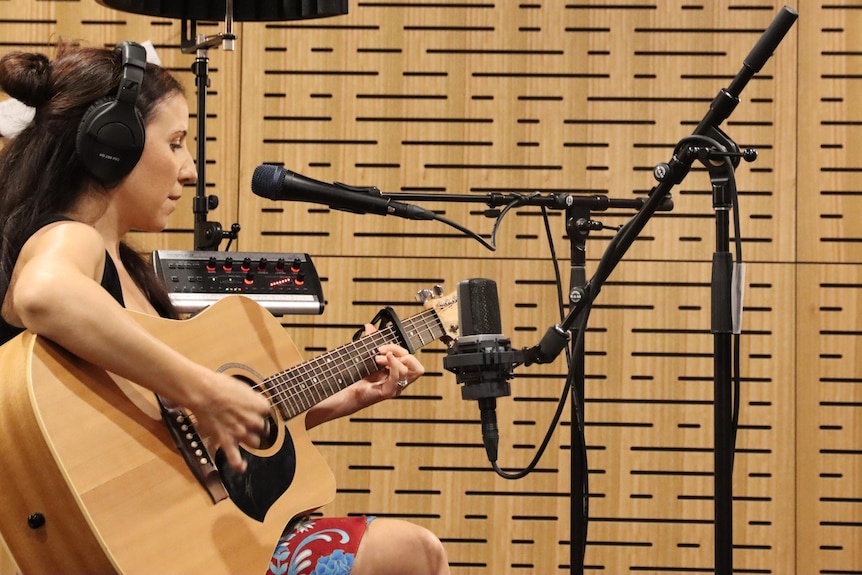 A side on picture of a woman playing guitar, with microphones set up in front of her.
