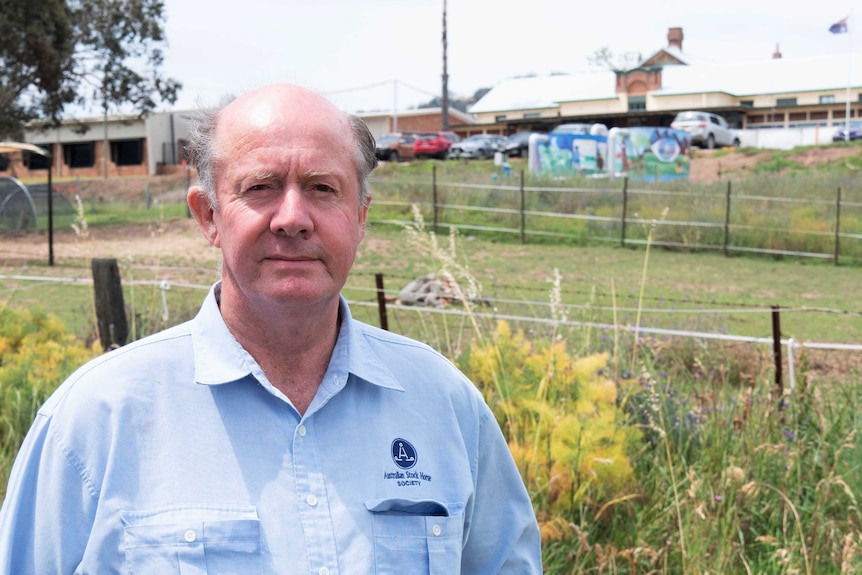 An older gentlemen in a light blue shirt standing, with an old looking building in the background.