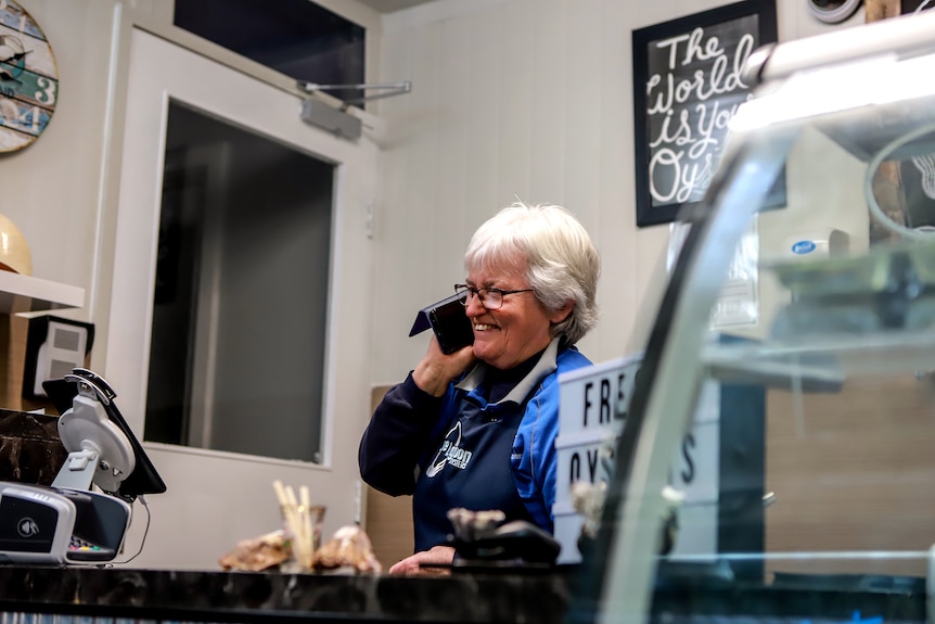 Woman with white hair wearing navy blue apron and blue shirt stands at shop counter holding phone