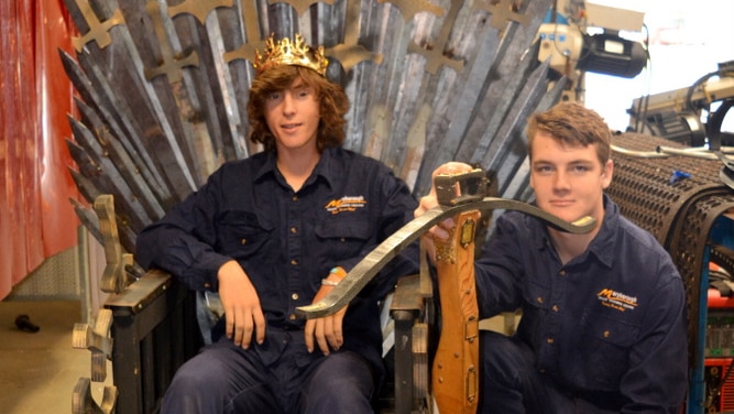 Students dressed on blue overalls sit on a replica of the Iron Throne.