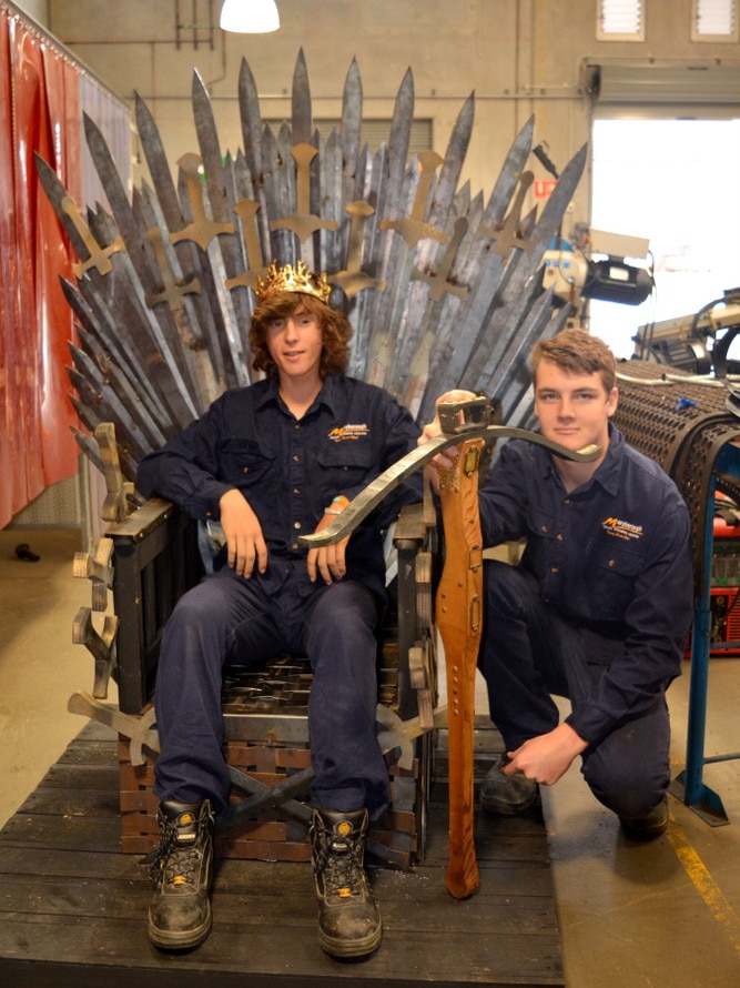 Students dressed on blue overalls sit on a replica of the Iron Throne.