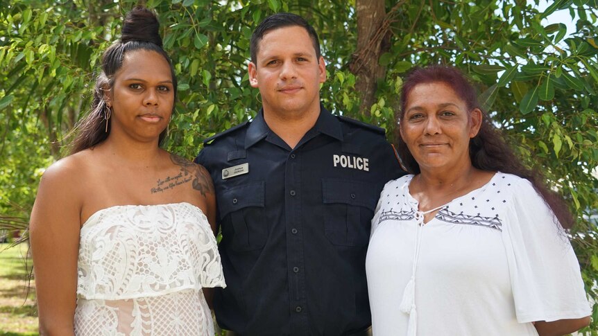 A police officer stands smiling with his partner and mother.