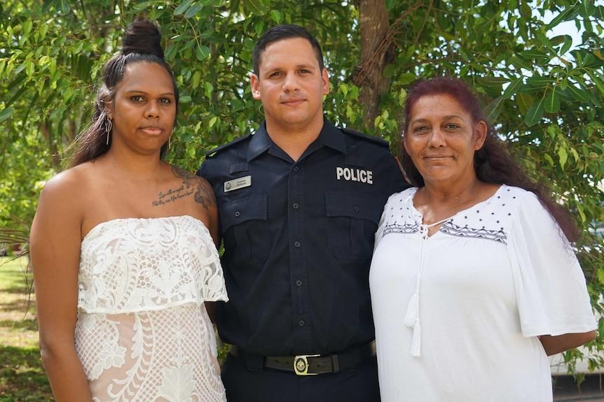 A police officer stands smiling with his partner and mother.