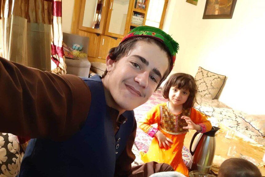 A boy in traditional Afghan hat taking selfie with girl in the background.