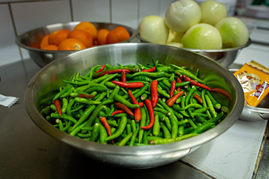 Three stainless steel bowls filled with fresh chillies, onions and tomatoes sit on a bench.